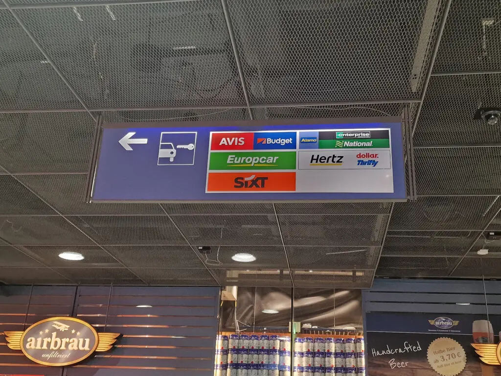 Rental car directional sign at Munich Airport showing Avis, Budget, Hertz, Sixt, Europcar and other agencies, with Airbrau beer shop visible below