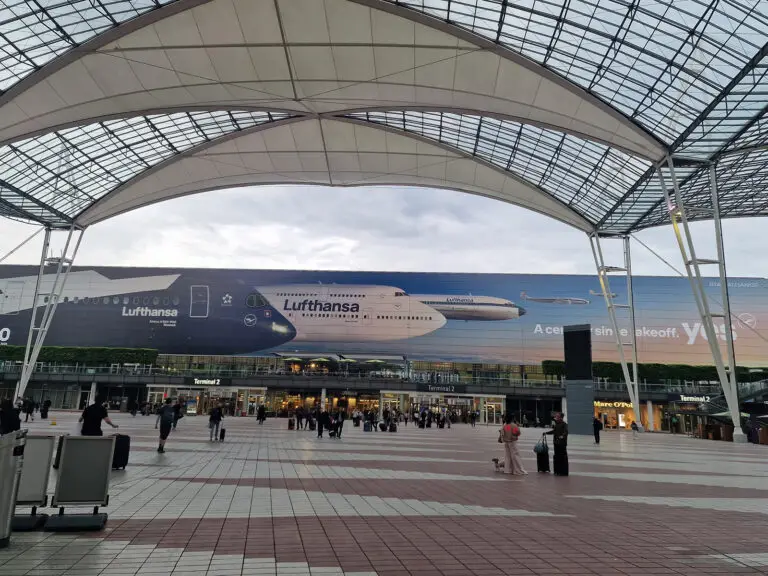 3:09 PM Terminal 2 forecourt at Munich Airport under a white canopy roof, with a large Lufthansa aircraft mural covering the terminal facade