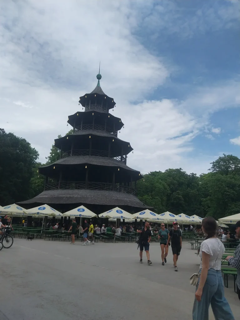 Chinesischer Turm beer garden in Munich's English Garden on a summer afternoon with outdoor tables, Hofbräu umbrellas, visitors, and a cloudy blue sky